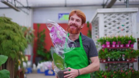 Foto de Feliz joven empleado de la tienda de plantas llevando flores. Un hombre pelirrojo caucásico vistiendo delantal verde camina por el pasillo de la tienda de negocios - Imagen libre de derechos