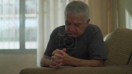 Téléchargez les photos : Homme âgé dévoué faisant le signe de la croix à la maison, homme caucasien âgé engagé dans la prière sur le canapé, incarnant la spiritualité et l'espoir - en image libre de droit