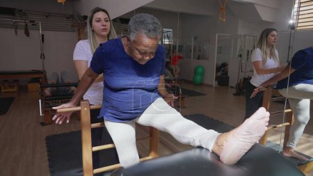Foto de Negro Senior Mujer estiramiento del cuerpo bajo Pilates Coach Instrucción, Participación en la sesión de rehabilitación de fisioterapia - Imagen libre de derechos