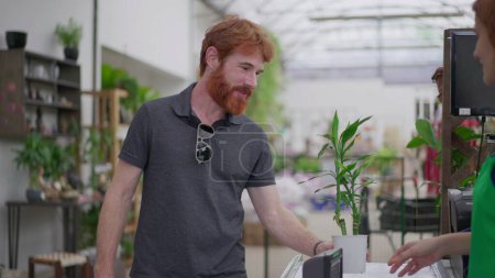 Photo for Young Man Purchasing Plant and Using Phone for Tap-to-Pay at Flower Shop. Seamless Contactless Payment at Horticulture Store - Royalty Free Image