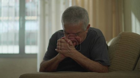 Téléchargez les photos : Homme âgé dévoué faisant le signe de la croix à la maison, homme caucasien âgé engagé dans la prière sur le canapé, incarnant la spiritualité et l'espoir - en image libre de droit