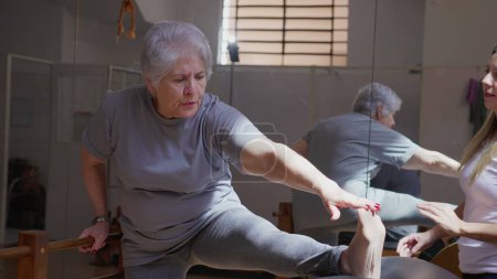 Foto de Entrenador de Pilates femenino guiando a una mujer mayor para estirar la pierna en clase de sesión de fisioterapia - Imagen libre de derechos