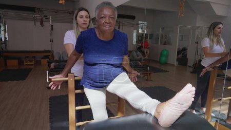 Téléchargez les photos : Femme âgée noire étirant le corps sous l'instruction d'entraîneur de Pilates, s'engageant dans la séance de réadaptation de physiothérapie - en image libre de droit