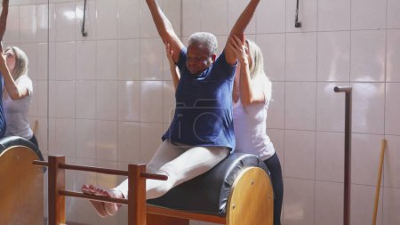 Photo for Female Pilates coach assisting an African American elderly woman to stretch body in Physiotherapy exercise session. Strength and flexibility old age workout lifestyle routine - Royalty Free Image