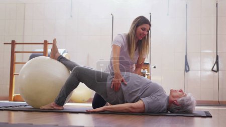 Téléchargez les photos : Physiothérapeute guidant une femme âgée dans un exercice de balle Pilates, dame âgée engagée dans une routine d'entraînement au sol avec l'aide d'un entraîneur - en image libre de droit