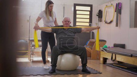 Photo for Senior man exercising with elastic bands seated on Pilates ball being oriented by a female coach instructor - Royalty Free Image