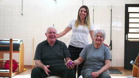 Photo for Senior man and woman sitting on Pilates Balls posing for camera with a young Sport Coach inside physiotherapist studio - Royalty Free Image