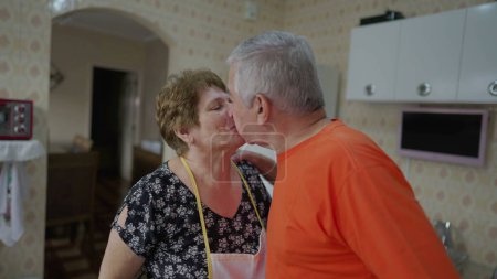 Photo for Older couple kissing in kitchen, wife kisses husband goodbye to start the day, senior couple peck kiss - Royalty Free Image