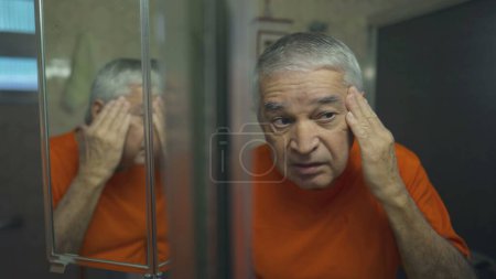 Téléchargez les photos : Homme âgé inspectant le visage et la peau, rituel de toilettage du matin reflété dans le miroir de salle de bains - en image libre de droit