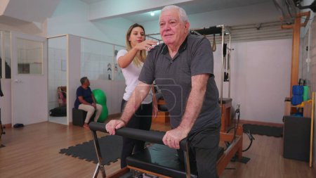 Téléchargez les photos : Homme plus âgé exerçant dans le studio Pilates étant orienté par entraîneur féminin. Personne âgée prenant soin de la santé de la colonne vertébrale - en image libre de droit