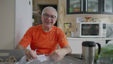 Photo for Senior man taking notes while seated in kitchen. A 70s male caucasian person writes information of paper with pen - Royalty Free Image