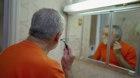 Foto de Hombre mayor afeitándose la barba delante del espejo del baño, la vejez estilo de vida doméstico ritual matutino - Imagen libre de derechos