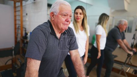 Téléchargez les photos : Homme âgé faisant de l'exercice dans le cadre d'un entraînement guidé, séance de Pilates Studio avec une entraîneuse, personne âgée utilisant une machine à étirer, prenant soin de la santé de la colonne vertébrale - en image libre de droit
