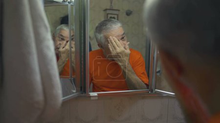 Téléchargez les photos : Homme âgé se toilettant devant le miroir pendant le rituel du matin. Style de vie domestique de la personne âgée dans les années 70 se préparer à inspecter le visage et la peau - en image libre de droit