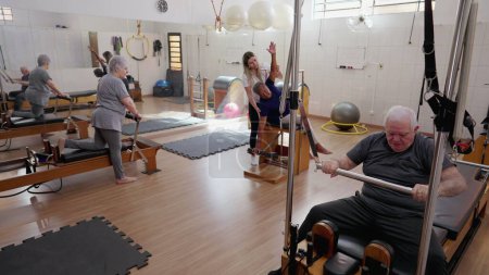 Photo for Elderly people exercising with Pilates Machines in Group Session. Female Coach Instructor guiding seniors to move and stretch body. Old age workout routine - Royalty Free Image