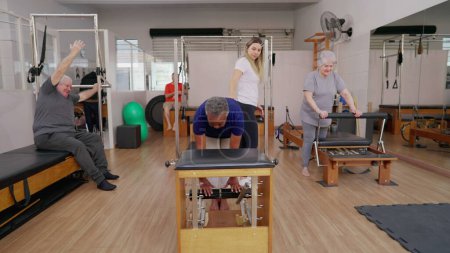 Photo for Group of elderly people doing Pilates Sessions with the help of female coach instructor. Older men and women using machines to stretch body and exercise, old age workout - Royalty Free Image