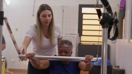Téléchargez les photos : Entraîneur de Pilates féminin aidant la femme âgée noire dans l'entraînement de machine pour le renforcement du corps et la flexibilité - en image libre de droit
