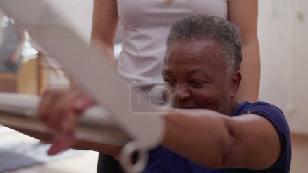 Téléchargez les photos : Entraîneur de sport féminin aidant une femme âgée afro-américaine noire à utiliser la machine Pilates, routine d'entraînement d'exercice à la vieillesse - en image libre de droit
