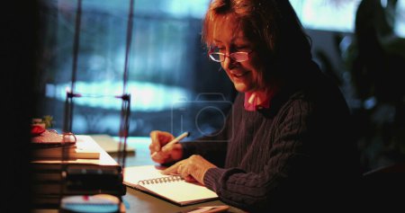 Photo for Older woman sitting at desk in evening writing letter - Royalty Free Image