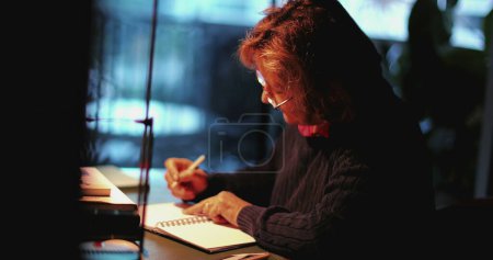 Téléchargez les photos : Femme âgée assise au bureau dans la soirée lettre d'écriture - en image libre de droit