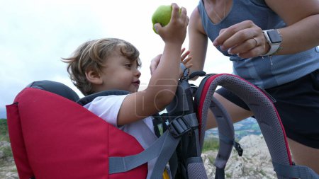 Photo for Child holding apple healthy snack during hike with mother inside carrying kid backpack - Royalty Free Image