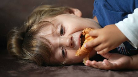 Photo for Child eating croissant one little boy lying on sofa eats carb food - Royalty Free Image