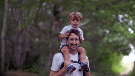 Téléchargez les photos : Père et fils marchant ensemble enfant sur les épaules du père regardant la caméra pendant la randonnée - en image libre de droit