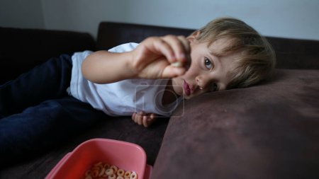 Photo for Child eating cereal little boy snacking lying on couch - Royalty Free Image