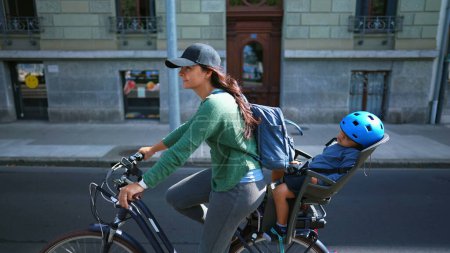 Téléchargez les photos : Femme heureuse à vélo avec enfant endormi sur le siège arrière dans la rue urbaine - en image libre de droit
