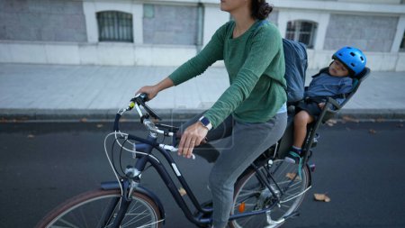 Téléchargez les photos : Cycliste mère équitation vélo dans la rue avec tout-petit sur le siège arrière - en image libre de droit