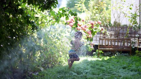 Foto de Niño pequeño que se divierte corriendo bajo la manguera de pulverización de agua en el jardín durante el día de verano - Imagen libre de derechos