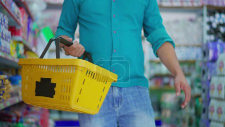 Téléchargez les photos : Gros plan de la main de customer _ s tenant un panier de supermarché, faisant du shopping pour des articles en marchant dans une allée d'épicerie - en image libre de droit