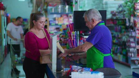 Foto de Escena de la tienda de comestibles de clientes mujeres que pagan por productos con teléfono en la caja del cajero en el supermercado interactuando con el personal de empleados senior - Imagen libre de derechos