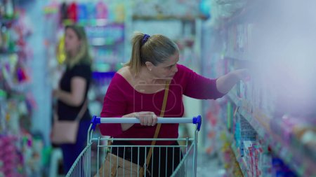 Foto de Compradora de comestibles hembra caminando en el pasillo buscando productos en el estante. Mujer de mediana edad mirando los productos en exhibición con carrito de compras - Imagen libre de derechos
