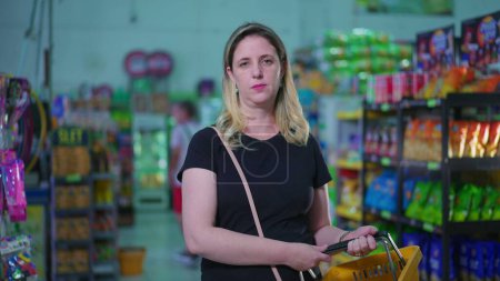 Téléchargez les photos : Consommateur féminin debout dans le panier de maintien de supermarché avec visage portrait d'expression grave gros plan dans le mouvement de tir de suivi - en image libre de droit