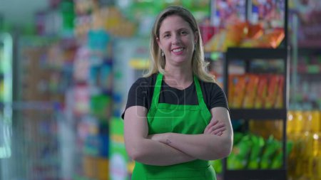 Téléchargez les photos : Joyeuse employée de l'épicerie avec les bras croisés souriant à l'intérieur de l'allée du supermarché, regardant la caméra, concept d'occupation d'emploi - en image libre de droit