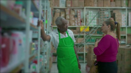 Téléchargez les photos : Un homme noir cadre supérieur à l'écoute de la cliente penché sur l'étagère et portant tablier vert, supermarché épicerie cadre de petite entreprise - en image libre de droit