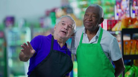Téléchargez les photos : Divers travailleurs âgés discutant des affaires de supermarché portant des tabliers. Un gestionnaire caucasien plus âgé expliquant le travail à un personnel brésilien noir - en image libre de droit