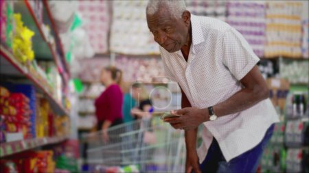 Foto de Un hombre negro senior comprando productos de pie en el pasillo de la tienda de comestibles navegando artículos mientras sostiene y mira el teléfono - Imagen libre de derechos