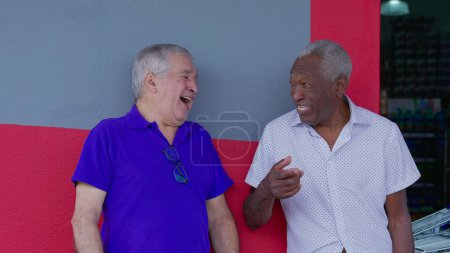 Photo for Joyful two diverse male senior friends smiling and laughing together leaning on sidewalk wall. A caucasian and African American older men interaction - Royalty Free Image