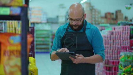 Foto de Supermercado personal de inventario de productos de navegación con dispositivo de tableta. Empleado masculino que usa delantal administrando artículos de la tienda de comestibles para la venta - Imagen libre de derechos