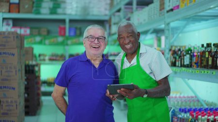 Photo for Two senior employees of Grocery store posing for camera smiling inside small business while holding tablet. Older diverse managers of supermarket - Royalty Free Image