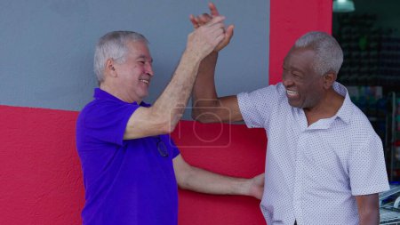Photo for Two happy diverse older friends hugging and celebrating with high-five standing outside on sidewalk. Cheerful African American and caucasian companionship authentic interaction - Royalty Free Image