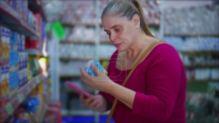 Téléchargez les photos : Female Shopper Cross-Checking Détails du produit En ligne Utilisation du smartphone à l'épicerie, illustrant l'utilisation de la technologie dans les achats - en image libre de droit