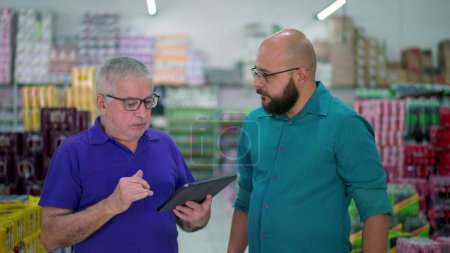 Photo for Senior manager of grocery store speaking with staff while looking at tablet device. Two men standing inside local business, job occupation delegation concept - Royalty Free Image