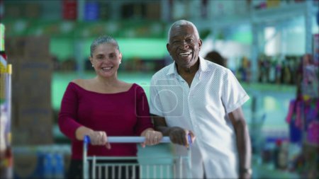 Téléchargez les photos : Joyeux clients de l'épicerie posant pour caméra debout à l'intérieur des affaires avec panier. Un vieil homme noir et une femme caucasienne d'âge moyen - en image libre de droit