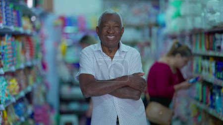 Téléchargez les photos : Un gestionnaire brésilien senior noir de l'épicerie tenant tablette posant pour appareil photo à l'intérieur de la petite entreprise. Une personne afro-américaine plus âgée utilisant la technologie moderne - en image libre de droit
