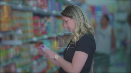 Téléchargez les photos : Épicerie féminine regardant l'article de l'étagère debout dans l'allée, femme lisant l'étiquette du produit - en image libre de droit