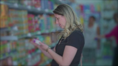 Photo for Female grocery shopping looking at item from shelf standing inside aisle, woman reading product label - Royalty Free Image