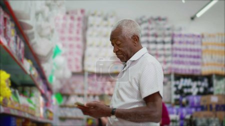 Téléchargez les photos : Un homme noir senior magasinant pour les produits debout à l'épicerie allée articles de navigation tout en tenant et en regardant le téléphone - en image libre de droit